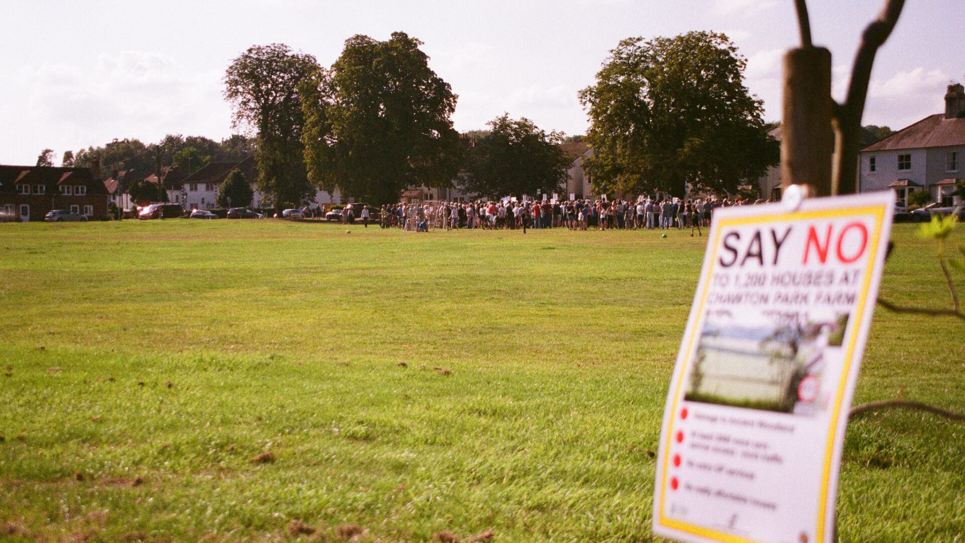 A campaign poster "say no to" with a protest in the distance, on a green field
