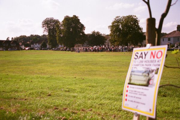 A campaign poster "say no to" with a protest in the distance, on a green field